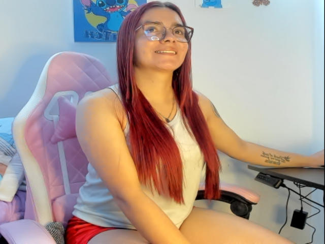 EimmyHoskX with red hair, glasses, white tank top, red shorts, sitting on pink and white gaming chair, smiling, tattoo on right arm, hands on desk.