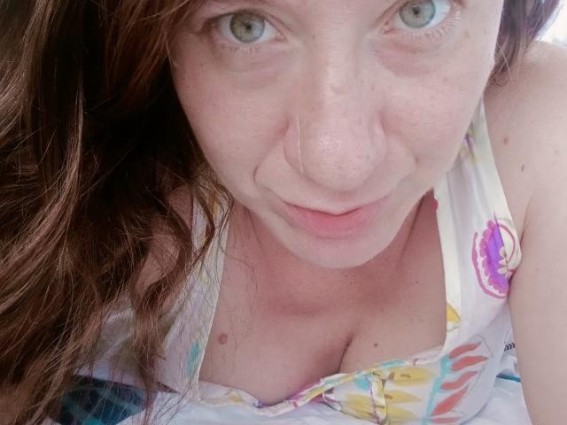 MissGinger's face is close to the camera, looking up with blue eyes and a slight smile. She has pale skin, brown wavy hair, and visible cleavage in a colorful top.