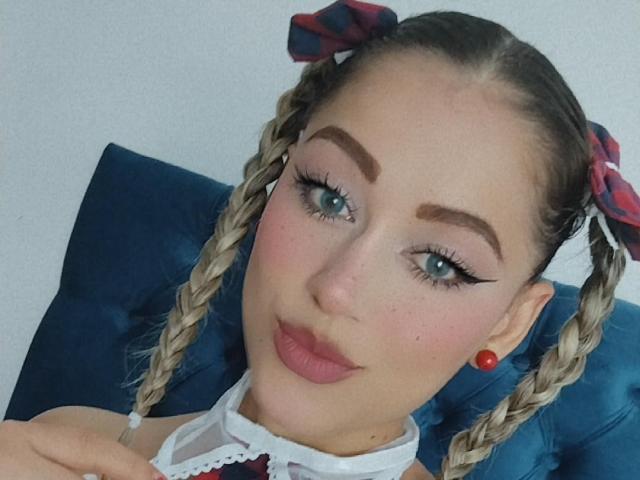 Samhanttha with braided blonde hair, plaid bows, red earrings, thick eyebrows, blue eyes, and pink lipstick, sitting on a blue cushion. Her tits are covered by a white top.