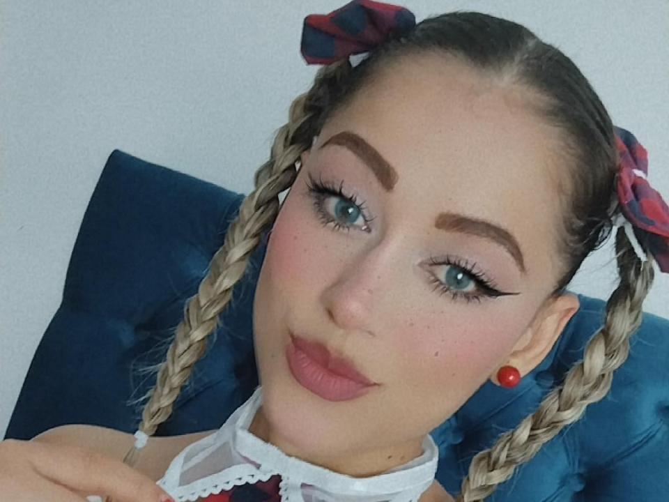 Samhanttha with braided blonde hair, plaid bows, red earrings, thick eyebrows, blue eyes, and pink lipstick, sitting on a blue cushion. Her tits are covered by a white top.