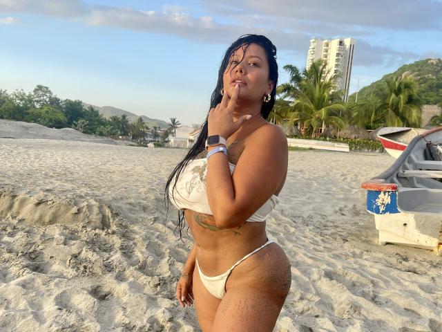 AlessandraMalory stands on a sandy beach, wearing a white bikini, her hand near her mouth, wet black hair, tan skin, curvy with big ass, and small white tits.