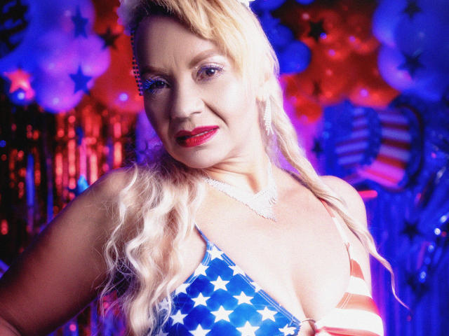 Anastasia Taylor, com cabelo loiro, batom vermelho e um colar brilhante, vestindo uma blusa azul e branca com estampas de estrelas e listras vermelhas, exibindo seus seios grandes, em um ambiente colorido e festivo..