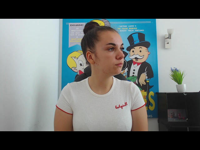 ReineDuJeu with puffy ass, white tee, red trim, and "Bella" in red, pouting, dark hair in bun, cartoon poster in background, black shelf with plant, white wall.