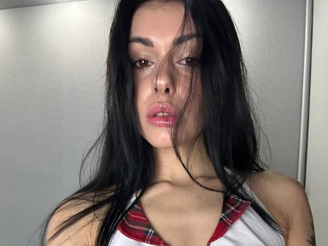 EmmyWarm with long black hair, pink glossy lips, and a red plaid collar on a white shirt, looking slightly to the side with a subtle, seductive expression.