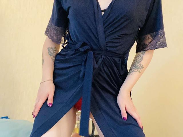 KarKutie in a navy blue satin robe with lace sleeves, hands on her thighs, showing red panties, tattoos on her arms, nail polish, and a ring on her left hand.