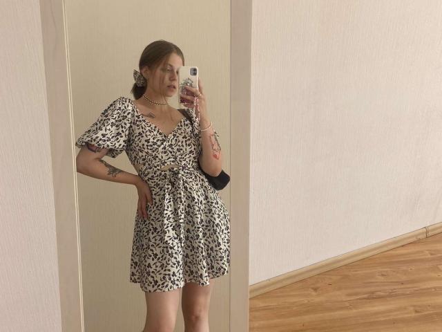 KarKutie with brown hair, wearing a leopard print dress, holding a phone, showing tattoos, standing in front of a mirror, wooden floor, white walls, hand on hip.