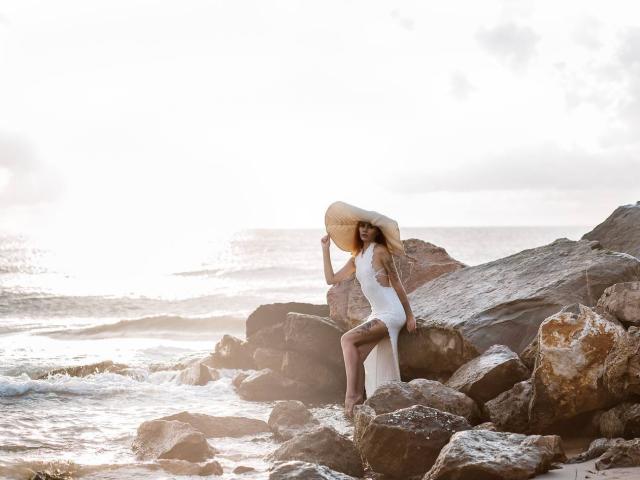 EmmyWarm with sunhat, white dress, and bare legs, sitting on rocks by the sea, holding hat, back to camera, ocean and sunlight in background, looking sexy.
