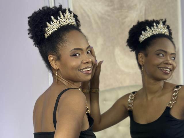 ChantalHaneke with dark skin, curly black hair, wearing a gold tiara and black dress, smiling at her reflection in a mirror. She's touching her cheek, showing her ass.