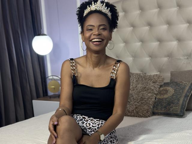 ChantalHaneke with dark skin, short curly hair, wearing a sparkly headpiece, black tank top with gold straps, leopard print skirt, large hoop earrings, smiling, sitting on a bed.