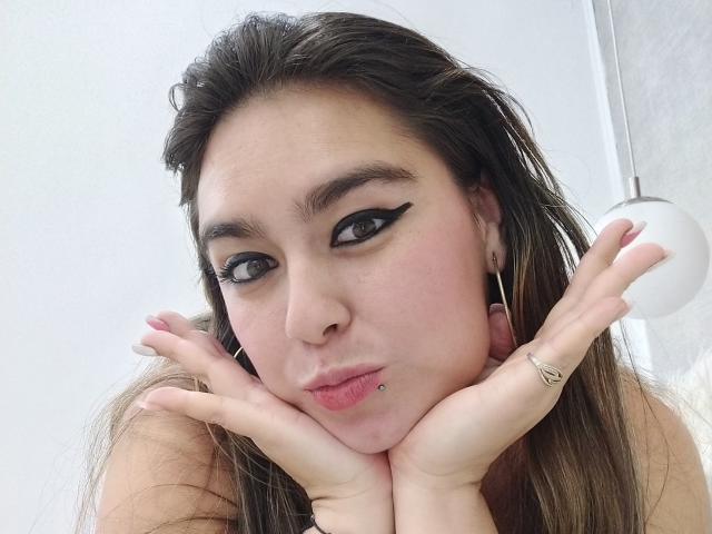 TaylorNova, avec des lèvres charnues, un trait d'eye-liner noir et ailé, et un piercing au nez, les mains sous le menton, des cheveux bruns, et une bague en argent à sa main droite, regardant la caméra..