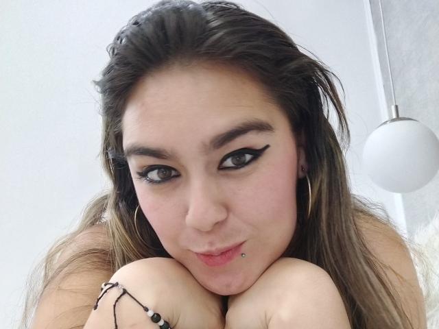 TaylorNova with black eyeliner, pouty lips, and a lip piercing, leaning on knees, wearing hoop earrings and a black bracelet, looking up with brown eyes.