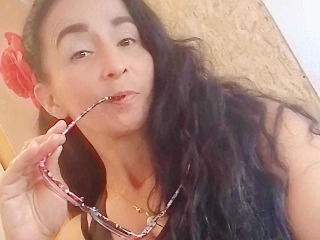 CristalVelvet with long black hair, holding pink-framed glasses in her mouth, wearing a black top and a gold necklace, has a red flower in her hair.