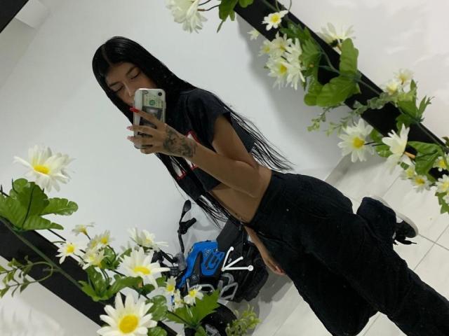 SweetestBebe with long black hair, small tits, and a tattoo on her left hand, wearing a black crop top and black pants, taking a mirror selfie with white flowers.