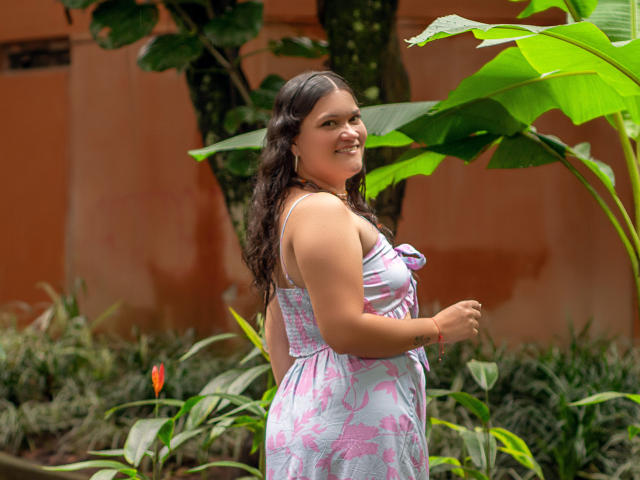 LizzieSin with curly brown hair, smiling, wearing a pink and blue floral dress, standing in a garden, showing her curvy ass, with green leaves and orange flower in background.