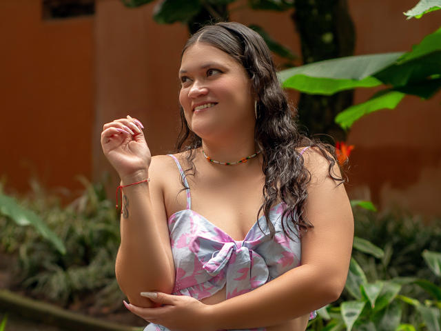 LizzieSin with wavy brown hair, smiling, wearing a pink and blue patterned top, showing small tits, arm raised, colorful necklace, green plants in background.