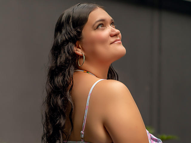LizzieSin with long wavy black hair, tan skin, and hoop earrings, looking up with a slight smile, showing her bare shoulders and pink halter top. Her tits are partially visible.