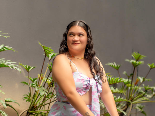 LizzieSin with long dark wavy hair, light pink floral bikini top, and matching bottom, standing among green plants, smiling, showing a tattoo on her left forearm.