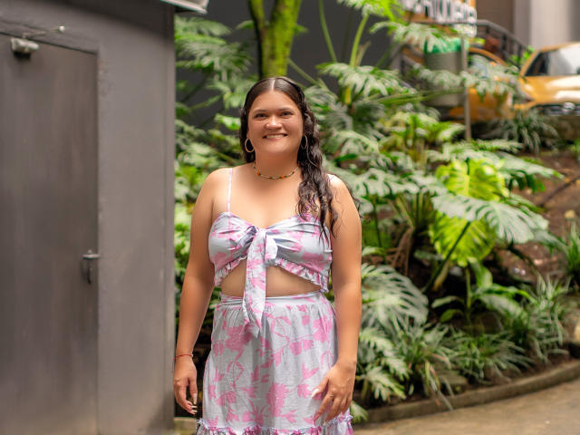 LizzieSin with long brown hair, smiling, wearing a pink and white patterned, tied-up top and matching skirt, standing by a gray wall, surrounded by green plants.