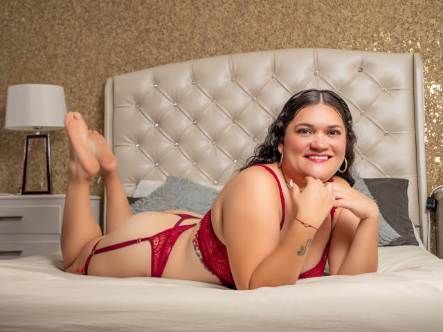 LizzieSin, a curvy woman with dark hair, lies on a bed in red lace lingerie, smiling with her ass raised and legs up, showing her tits and pussy.