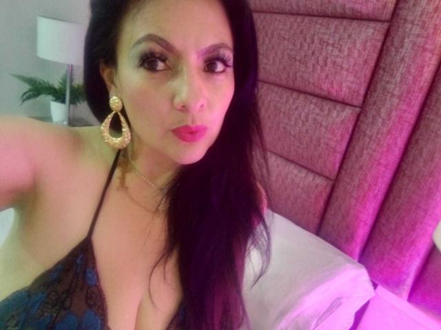 JakyKidman with long black hair, wearing gold hoop earrings and a blue lace dress, showing cleavage and pink lipstick, looking at the camera with a slight pout.