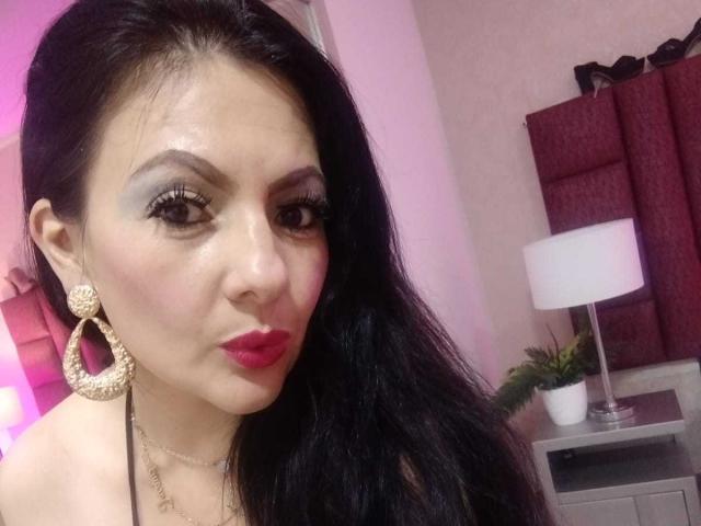 JakyKidman has dark hair, red lipstick, and large gold hoop earrings. She's wearing a necklace and has long eyelashes. Her tits are not visible in this shot.