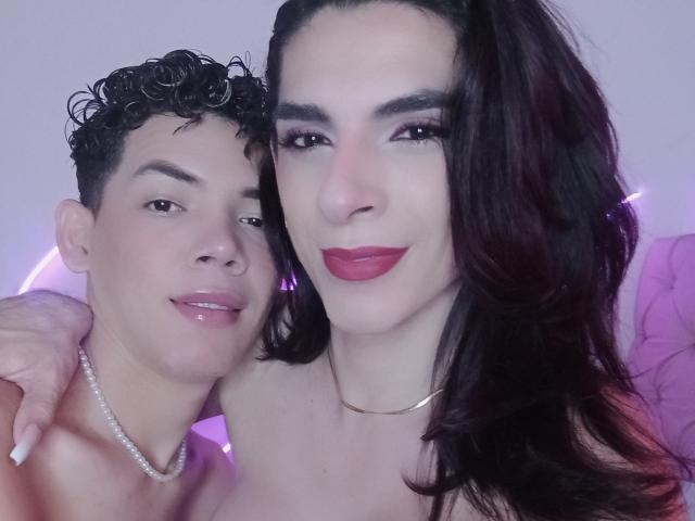 AdamPaulUnlimited with curly black hair and a necklace, standing next to AdamPaulUnlimited with red lipstick, long dark hair, and a gold choker, both smiling.