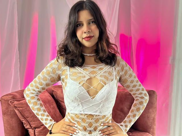 ScarlethSweetX with dark wavy hair, wearing a white fishnet top, exposing small tits, hands on hips, pink and white curtain, pink light, smiling, red velvet chair.