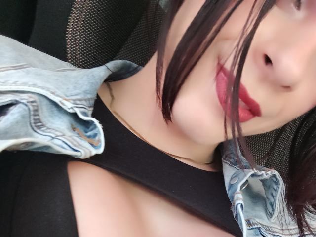 AdamPaulUnlimited with red lipstick, black top, and blue denim jacket, exposing cleavage, with black hair partially covering face. Soft focus, slight smile. Upper chest and face visible.