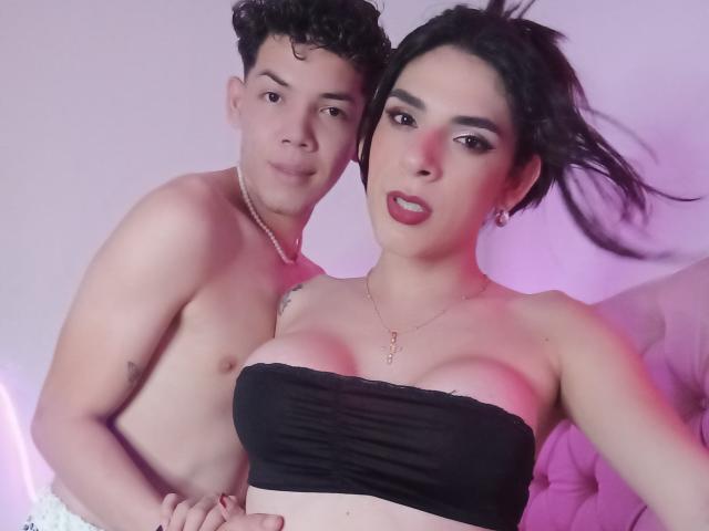 AdamPaulUnlimited with curly brown hair, shirtless, and AdamPaulUnlimited with black strapless top, showing large tits, and pink lipstick, standing closely, both with light skin and casual expressions.
