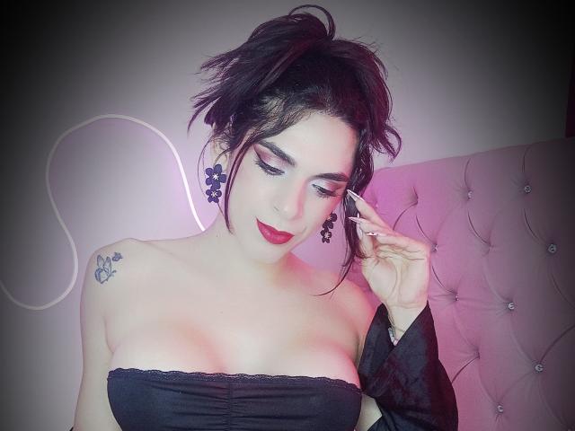 AdamPaulUnlimited with dark hair, red lipstick, black strapless top, showing tits, tattoo on left shoulder, adjusting ear, pink and purple lighting, looking down, black earrings.