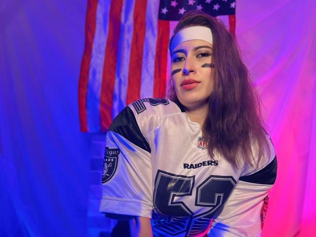 VidiaStons met lang bruin haar, die een witte hoofdband draagt, een American football jersey met het nummer 26, en gezichtsverf, onder blauw en roze verlichting. Amerikaanse vlag op de achtergrond..
