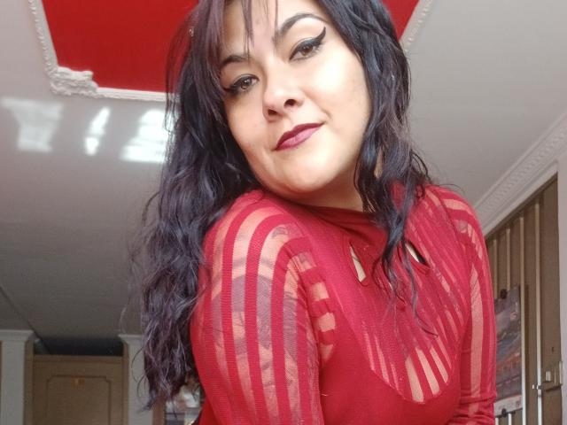 FreeMystic69 met donker, golvend haar, een rode top met strepen en donkere lipstick, glimlacht naar de camera en laat haar borsten zien, met een schattig gezicht. Haar eyeliner is scherp en ze straalt zelfvertrouwen uit..