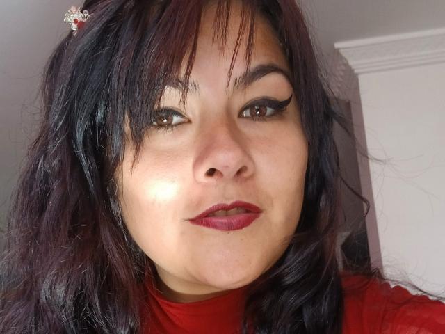 FreeMystic69 heeft donker, golvend haar, bruine ogen met dikke zwarte eyeliner, rode lippenstift en een rode top. Ze glimlacht en haar gezicht wordt zacht verlicht..