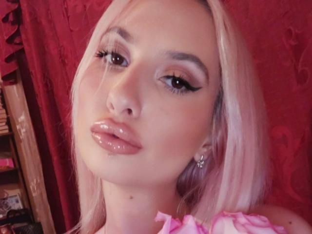 ElzaKomx has shiny pink lips, long eyelashes, and platinum blonde hair. She's wearing pink ruffled top, with a small mole on her neck. Red background.