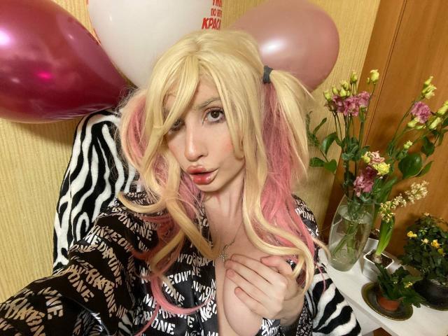 Blonde ElzaKomx with pink pigtails, pouty lips, and zebra-pattern robe, holding her tits, surrounded by pink and white balloons, in a room with flowers.
