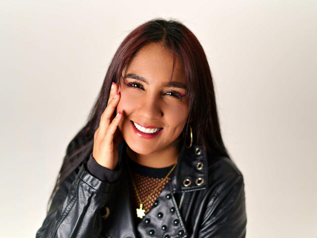 GipsyRouse with dark red hair, smiling, wearing a black leather jacket with studs, gold hoop earrings, and a fishnet top, touching her cheek with red-painted nails.