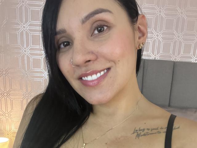 YessiJaymes has black hair, a nose ring, and a gold necklace. She's smiling with white teeth, showing a tattoo on her right shoulder and gold earrings.