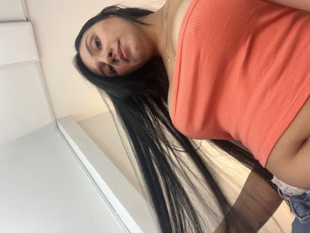 YessiJaymes, con lunghi capelli neri, indossa un top a tubino arancione a coste, che mette in mostra i suoi piccoli seni, è in piedi con il sedere leggermente scoperto, indossa biancheria intima in pizzo, e guarda la fotocamera..