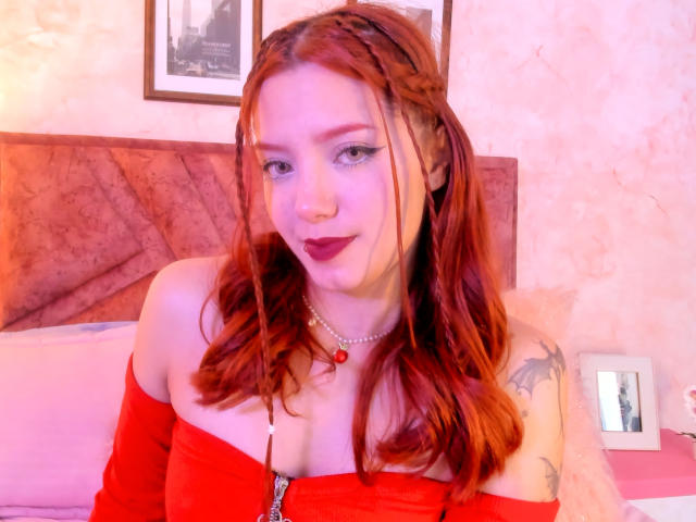 EmmaThomsonn with red hair, braided, off-shoulder red top, showing her tits, dark lipstick, nose ring, necklace, tattoo on right shoulder, smiling, pink and purple lighting.