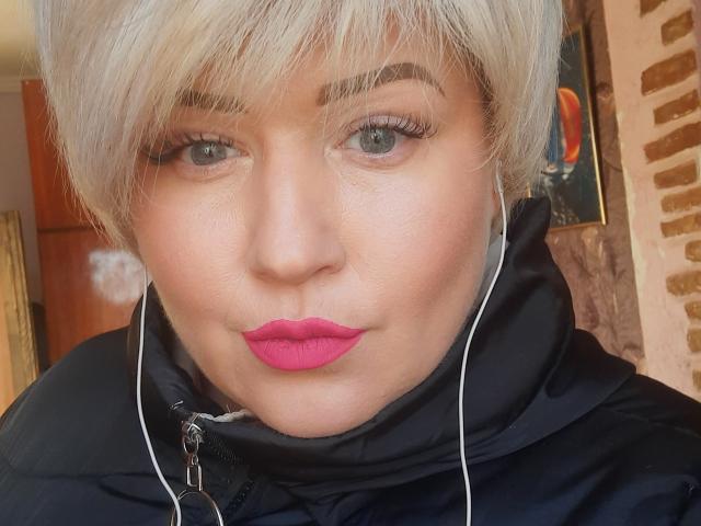 A RadaFoxy tem cabelo loiro curto, batom rosa e está vestindo uma jaqueta preta com fones de ouvido, olhando para a câmera com um leve semblante de descontentamento..