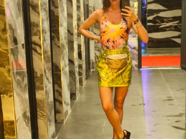 LucieDeluxe, atractiva con pequeños senos, vistiendo una blusa colorida con un estampado de sol, una falda amarilla brillante con un cinturón blanco, y sandalias negras, tomándose un selfie frente al espejo..