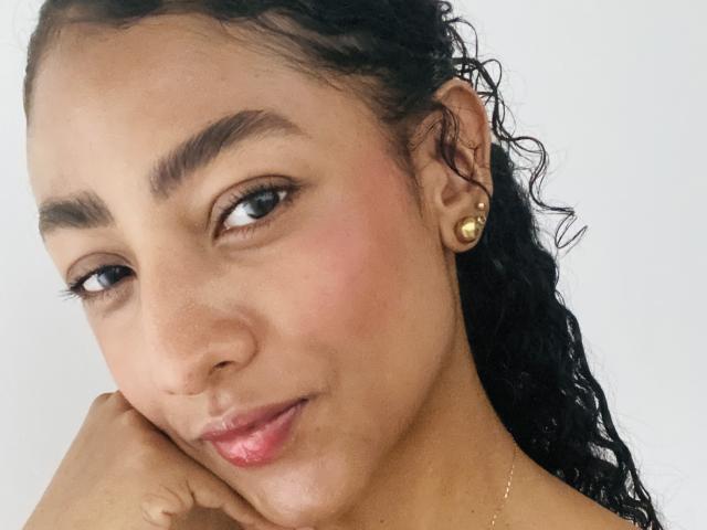 NatalyBlonder has curly black hair, brown eyes, and a cute face with pink blushed cheeks. She's wearing gold earrings and slightly pouting with her hand on her chin.