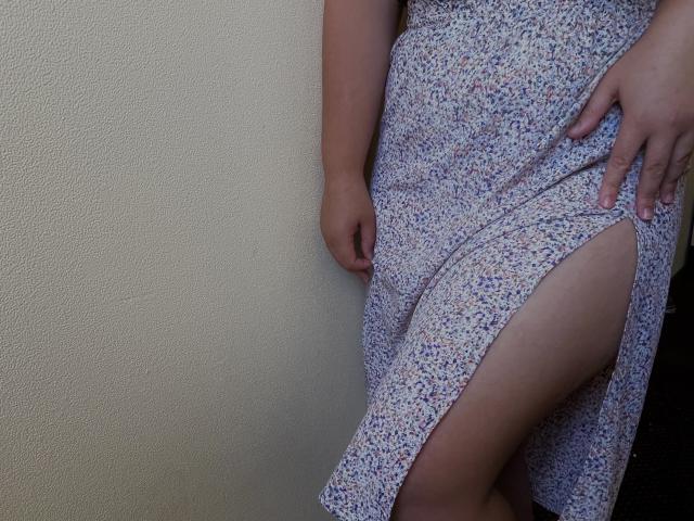 La robe fleurie de Madrinia, avec une fente haute sur le côté, révèle sa cuisse pâle. Elle est appuyée contre un mur texturé, sa main reposant sur sa hanche. Sa robe est légèrement relevée..
