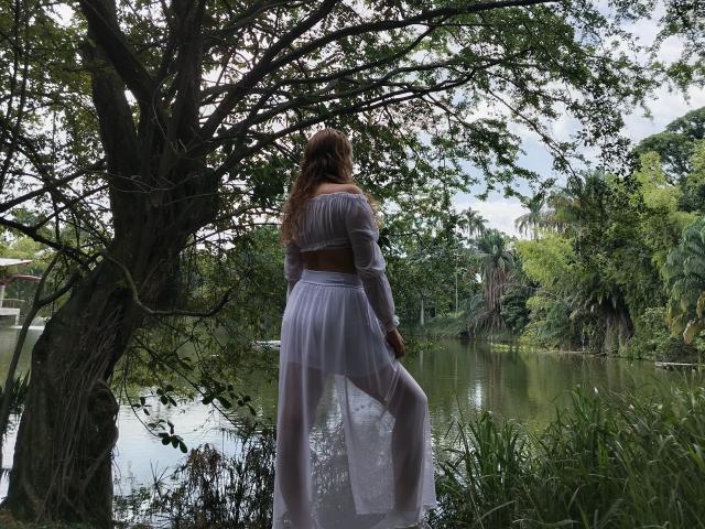 KeiliSexy stands with her back to the camera, wearing a sheer white top and long skirt, her ass slightly raised, brown hair flowing, by a green, tree-lined pond.