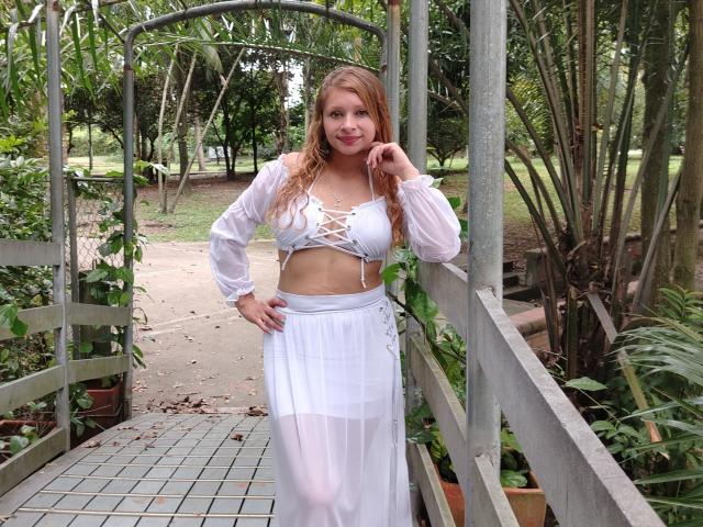 KeiliSexy with long blonde hair, white lace-up crop top, and sheer white skirt, standing on a tiled path, hand on hip, under a green archway, smiling.