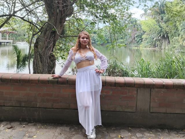 KeiliSexy with long red hair, white sheer crop top, and white flowing skirt, leaning on a brick wall, showing her tits and white sneakers, standing by a calm river.