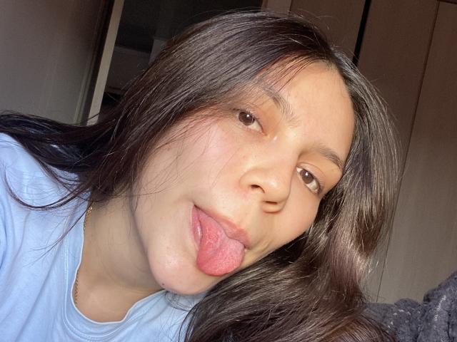 SophiaMarx with long brown hair, light skin, and brown eyes, sticking out her pink tongue in a selfie, wearing a light blue shirt. Her face is centered and close-up.