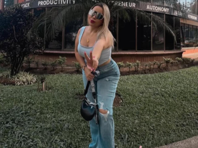 MiaBlondeHot with blue ripped jeans, grey tank top, holding black bag, standing on grass, wearing sunglasses, blonde hair, large tits, curvy ass, smiling, reaching forward.