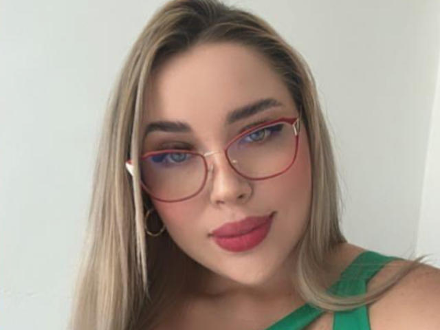 MiaBlondeHot has blue eyes, red glasses, and red lipstick. She's wearing a green top with long blonde hair, and gold hoop earrings, looking cute and confident.