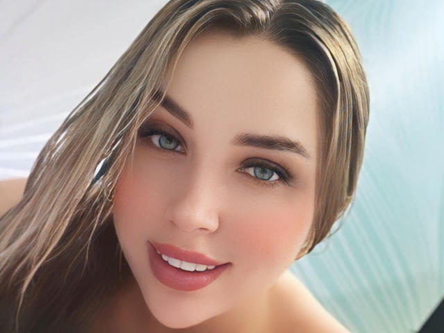 MiaBlondeHot has blue eyes, full pink lips, and a cute smile. Her blonde hair is wet, and she's looking directly at the camera with a sexy, relaxed expression.